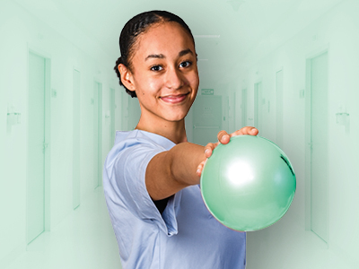 Lächelnde junge Frau in hellblauem Shirt streckt einen grünen Ball nach vorn. Heller Flur im Hintergrund; Fokus auf aktive, freundliche Geste.