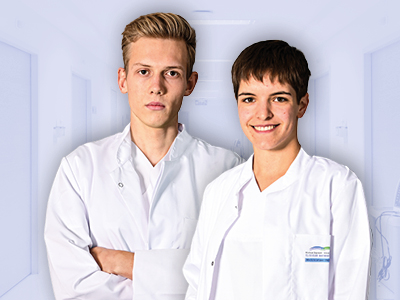 Zwei junge Mitglieder des medizinischen Personals in weißen Kitteln stehen im Krankenhausflur; links ernst, rechts lächelnd. Das Bild vermittelt Professionalität, Kompetenz und Bereitschaft zur Versorgung.